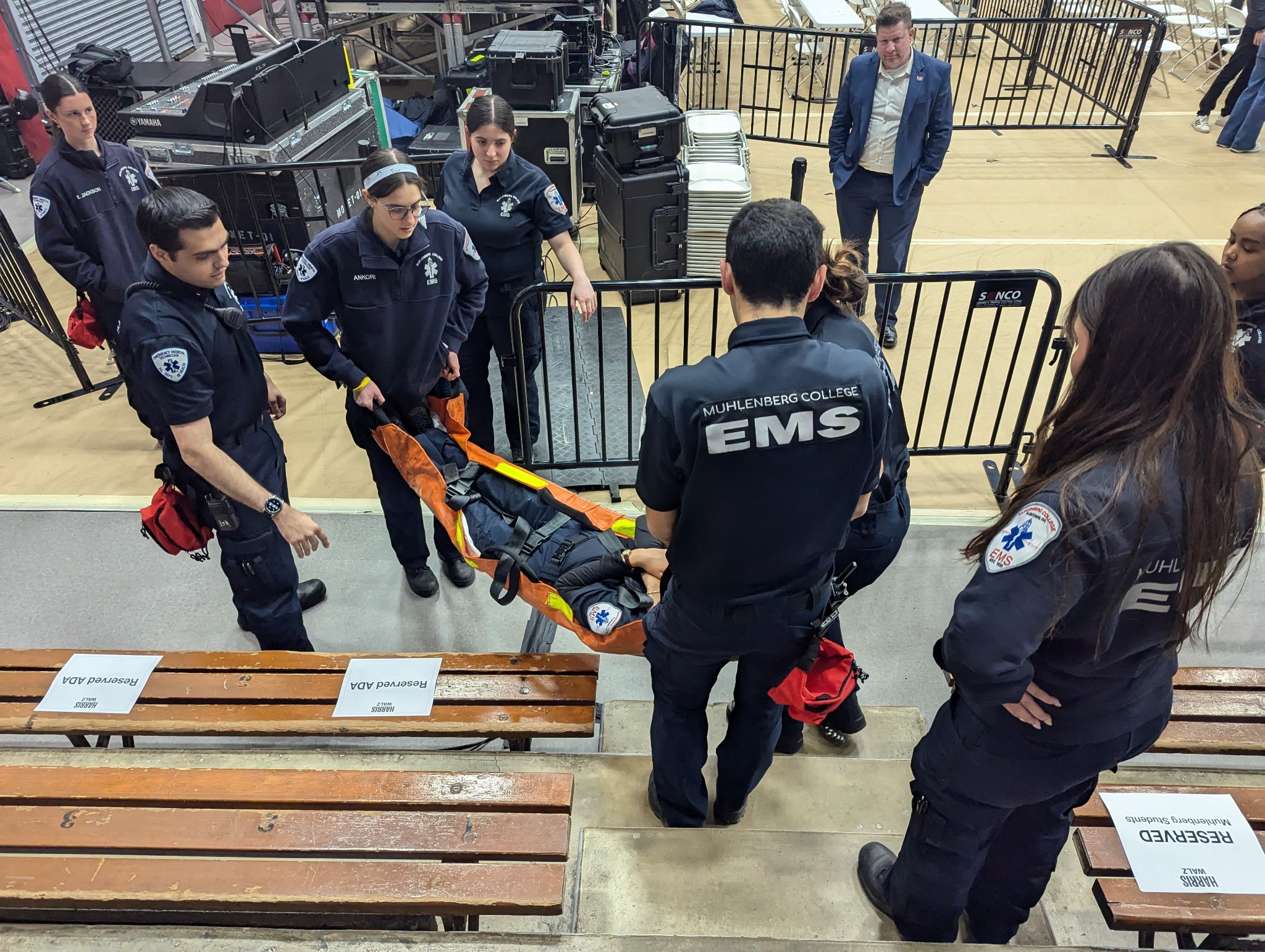 A group of college students in EMS uniforms carries a stretcher down a set of bleachers in a gymnasium